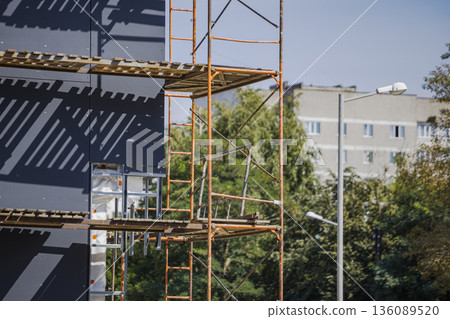 Workers are using scaffolding to renovate a building next to other structures in a busy area under a clear blue sky in the afternoon light Workers are using scaffolding to renovate a building next to other structures in a busy area under a clear blue sky in the afternoon light 136089520