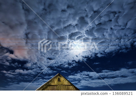 Bright moon illuminates clouds in the night sky above a barn in a rural area. Power lines run across the scene, adding to the landscape Bright moon illuminates clouds in the night sky above a barn in a rural area. Power lines run across the scene, adding to the landscape 136089521