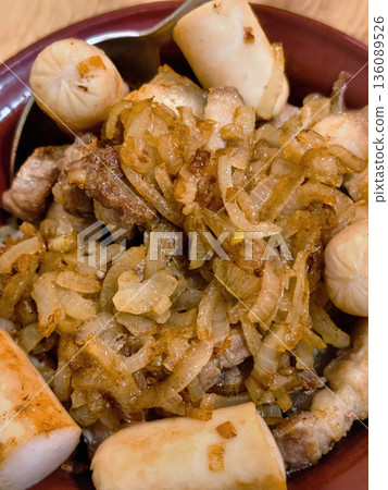 Slices of grilled meat with cooked onions and small sausages are served in a bowl on a dining table in the evening setting Slices of grilled meat with cooked onions and small sausages are served in a bowl on a dining table in the evening setting 136089526