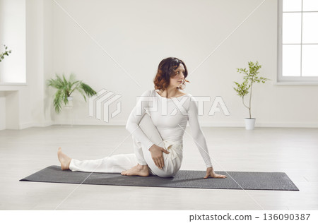 Woman in white sportswear doing warming up stretching exercises sitting on yoga mat indoors. 136090387