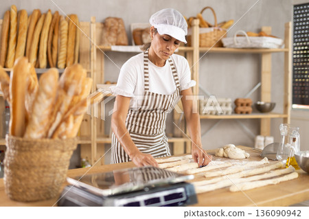 Female baker prepares raw yeast dough in bakery, forming it into long sausages to be cut into pieces for baking buns Female baker prepares raw yeast dough in bakery, forming it into long sausages to be cut into pieces for baking buns 136090942
