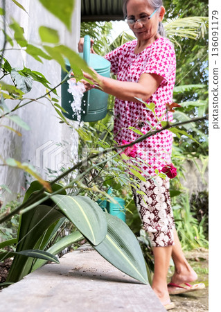 An elderly Filipina woman waters plants in the yard of her home An elderly Filipina woman waters plants in the yard of her home 136091179