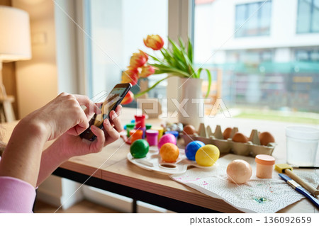 Woman taking photo of colorful Easter eggs with smartphone at home 136092659