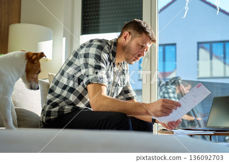 Man reviewing household bills on sofa with dog at home Man reviewing household bills on sofa with dog at home 136092703