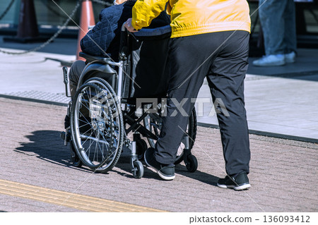 Yokohama cityscape in Japan. People wearing masks... An aging society. An elderly woman in a wheelchair wearing a mask... The person pushing the wheelchair on the right is also wearing a mask... Yokohama cityscape in Japan. People wearing masks... An aging society. An elderly woman in a wheelchair wearing a mask... The person pushing the wheelchair on the right is also wearing a mask... 136093412