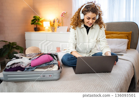 Young woman using laptop while packing suitcase for travel at home 136093501
