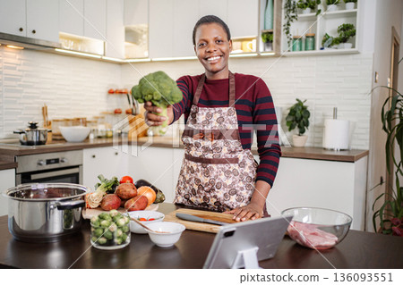 Woman holding broccoli cooking healthy meal guided by tablet Woman holding broccoli cooking healthy meal guided by tablet 136093551