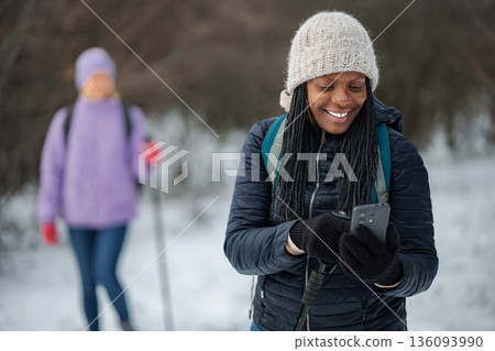 Woman smiling using smartphone while winter hiking with friend Woman smiling using smartphone while winter hiking with friend 136093990