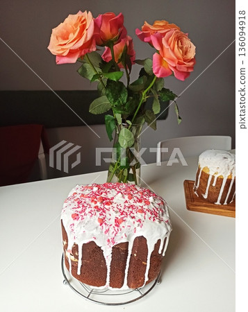 Easter cake with white icing and pink sprinkles placed on a table with roses in a vase. image represents holiday celebration home baking and festive tradition 136094918