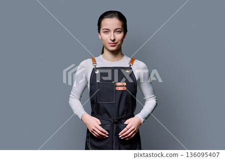 Portrait of young smiling female student in apron on gray background Portrait of young smiling female student in apron on gray background 136095407