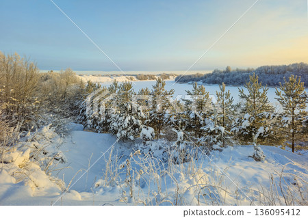 A snow-covered landscape with trees and a frozen river A snow-covered landscape with trees and a frozen river 136095412