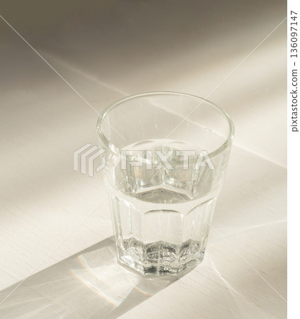 Glass of clean, fresh water on a white table, reflecting sunlight in the clear liquid, in a minimalist scene. Healthy lifestyle and hydration concept 136097147