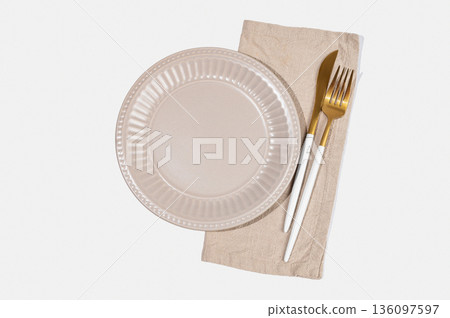 A ceramic beige plate with a linen napkin and cutlery on a light isolated background. A ceramic beige plate with a linen napkin and cutlery on a light isolated background. 136097597