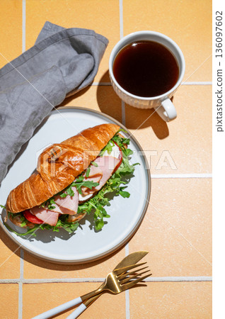 Croissant sandwich with ham, cheese, tomato and arugula on a plate on a yellow tile background Croissant sandwich with ham, cheese, tomato and arugula on a plate on a yellow tile background 136097602