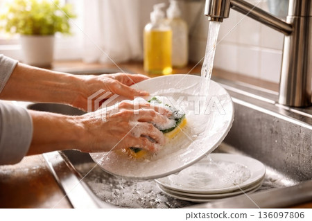 Scrubbing a Dirty Plate with Sponge Under Running Water 136097806