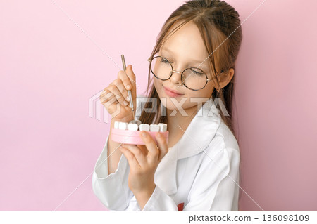 Child dressed as dentist examining teeth model with dental tools on pink background. Concept of pediatric dentistry, education, oral hygiene, healthy teeth, prevention and children healthcare. Child dressed as dentist examining teeth model with dental tools on pink background. Concept of pediatric dentistry, education, oral hygiene, healthy teeth, prevention and children healthcare. 136098109