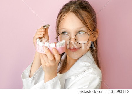 Child dressed as dentist holding teeth model on pink background. Concept of World Oral Health Day, oral hygiene awareness, prevention, healthy teeth education and children dental healthcare. Child dressed as dentist holding teeth model on pink background. Concept of World Oral Health Day, oral hygiene awareness, prevention, healthy teeth education and children dental healthcare. 136098110