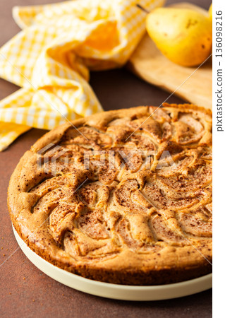 A fresh pear pie is placed on a cloth on a table. Nearby, a bowl holds ripe pears and a small vase has flowers. The setting looks warm and inviting for sharing a treat 136098216