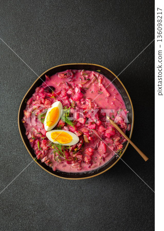 Delicious cold beet soup is garnished with boiled eggs and fresh herbs in a simple black bowl. The vibrant colors and textures create an inviting dish perfect for summer meals 136098217