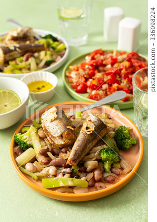 Breakfast includes baked hake placed on a bed of boiled beans with broccoli and peppers, accompanied by water and tomato salad on the table 136098224