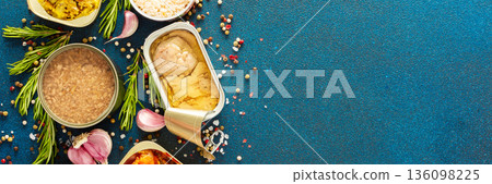 A top view of open tin cans with canned tuna, mackerel, shrimp, sardine, tuna, Cod liver, surrounded by spices and herbs on a blue background, banner 136098225
