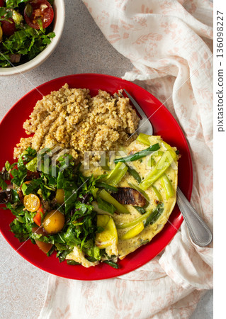 Grains, cooked greens, and vegetables are arranged on a red plate. A bowl of mixed salad with tomatoes is placed nearby on a light surface 136098227