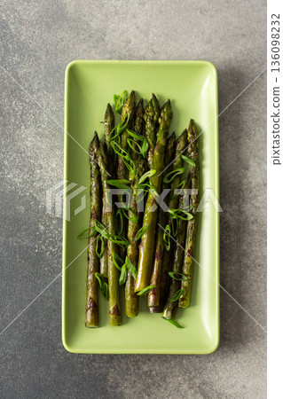 Asparagus spears are arranged neatly on a green plate, garnished with sliced green onions. This dish represents a simple yet flavorful vegetable choice for any meal Asparagus spears are arranged neatly on a green plate, garnished with sliced green onions. This dish represents a simple yet flavorful vegetable choice for any meal 136098232