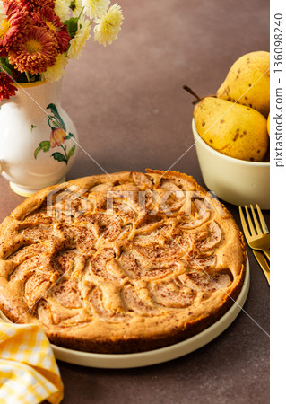 A fresh pear pie is placed on a cloth on a table. Nearby, a bowl holds ripe pears and a small vase has flowers. The setting looks warm and inviting for sharing a treat 136098240