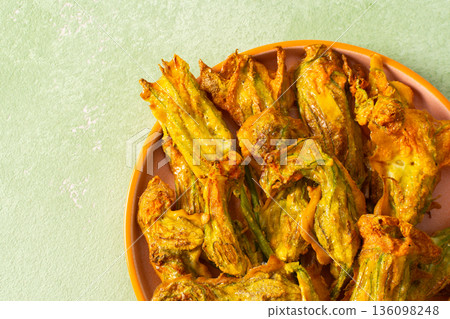 Crispy deep-fried zucchini flowers are displayed on a vibrant plate, showcasing their golden texture. Perfect for sharing during a summer gathering or family meal 136098248