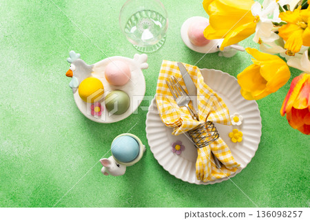 A beautifully arranged Easter table setting showcases a decorative egg nestled in a yellow napkin, surrounded by a glass of water and fresh flowers, perfect for holiday gatherings 136098257