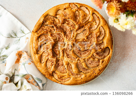 A fresh pear pie is placed on a cloth on a table. Nearby, a bowl holds ripe pears and a small vase has flowers. The setting looks warm and inviting for sharing a treat 136098266