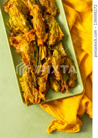 Delicious deep-fried zucchini flowers are neatly arranged on a green platter, showcasing their crispy texture. A bright yellow napkin adds warmth to the scene Delicious deep-fried zucchini flowers are neatly arranged on a green platter, showcasing their crispy texture. A bright yellow napkin adds warmth to the scene 136098272