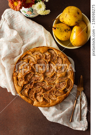 A fresh pear pie is placed on a cloth on a table. Nearby, a bowl holds ripe pears and a small vase has flowers. The setting looks warm and inviting for sharing a treat 136098275