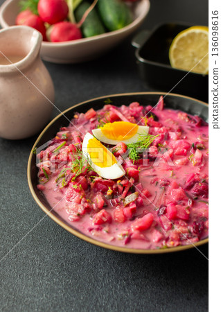 A vibrant bowl of cold beetroot soup features hard-boiled eggs and fresh herbs. A lemon slice accompanies it, all set against a black surface with a pink cloth for contrast A vibrant bowl of cold beetroot soup features hard-boiled eggs and fresh herbs. A lemon slice accompanies it, all set against a black surface with a pink cloth for contrast 136098616