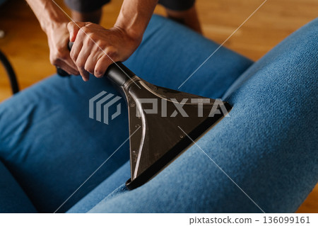 Close-up cropped shot of unrecognizable cleaning service worker using wet vacuum cleaner to remove dirt and stains from blue sofa, ensuring hygienic cleaning process. Concept of housekeeping 136099161