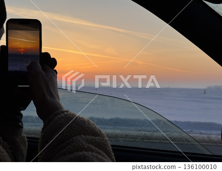 A driver of a passenger car has slightly opened the car window and is filming the sunset with a smartphone. A snow-covered field is visible to the horizon. Above it, a red solar disc and the glow of t A driver of a passenger car has slightly opened the car window and is filming the sunset with a smartphone. A snow-covered field is visible to the horizon. Above it, a red solar disc and the glow of t 136100010