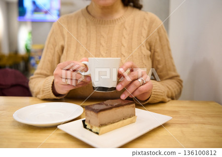 Woman In Beige Knit Sweater Sips Coffee Beside Chocolate Layer Cake Dessert At Table 136100812