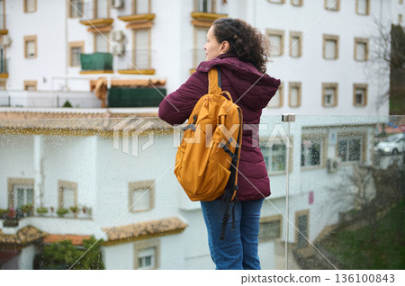 Woman With Orange Backpack Standing on Rainy Balcony Looking Over Cityscape 136100843