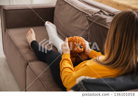 Young girl with broken leg cast sitting on sofa. Sad little kid hugging her teddy bear recovering from broken leg at home. Girl using cast and crutches. Accident injury fracture treatment foot bandage Young girl with broken leg cast sitting on sofa. Sad little kid hugging her teddy bear recovering from broken leg at home. Girl using cast and crutches. Accident injury fracture treatment foot bandage 136101058