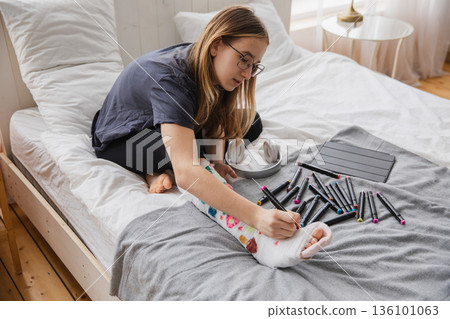 Young teenage girl with broken leg sitting on bed draws with felt tip pens on orthopedic cast. Bored girl with broken leg playing at home during rehabilitation. Accident injury fracture treatment 136101063