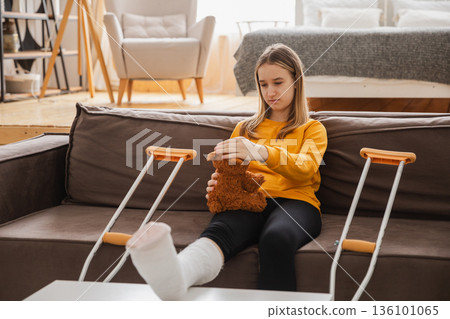 Young girl with broken leg cast sitting on sofa. Sad little kid hugging her teddy bear recovering from broken leg at home. Girl using cast and crutches. Accident injury fracture treatment foot bandage Young girl with broken leg cast sitting on sofa. Sad little kid hugging her teddy bear recovering from broken leg at home. Girl using cast and crutches. Accident injury fracture treatment foot bandage 136101065