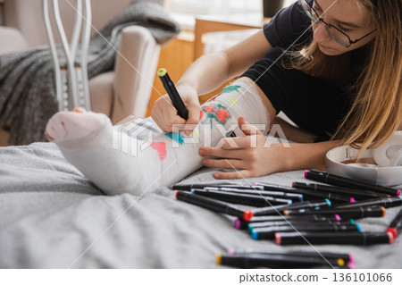 Young teenage girl with broken leg sitting on bed draws with felt tip pens on orthopedic cast. Bored girl with broken leg playing at home during rehabilitation. Accident injury fracture treatment Young teenage girl with broken leg sitting on bed draws with felt tip pens on orthopedic cast. Bored girl with broken leg playing at home during rehabilitation. Accident injury fracture treatment 136101066