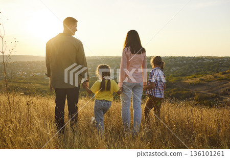 Family holding hands in golden meadow at sunset, looking over scenic valley 136101261