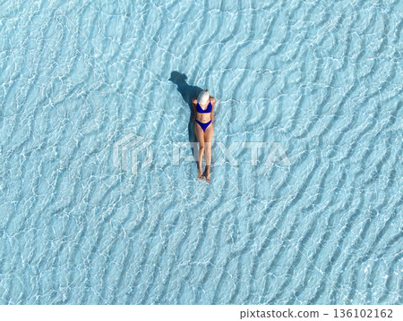 Aerial view of woman in bikini and white hat lying in blue sea Aerial view of woman in bikini and white hat lying in blue sea 136102162