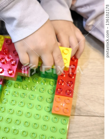 A toddler, sitting on the floor, builds a house out of bright construction sets on a green platform. A toddler, sitting on the floor, builds a house out of bright construction sets on a green platform. 136102178