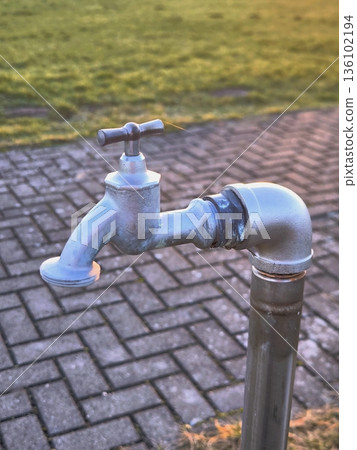 A street metal water tap against the backdrop of paving stones in a park A street metal water tap against the backdrop of paving stones in a park 136102194