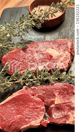 Preparing Flavorful Raw Steaks With Herbs On Rustic Kitchen Surface, Freshly Cut Raw Steaks Garnished With Thyme And Rosemary On Traditional Wooden Cutting Board Preparing Flavorful Raw Steaks With Herbs On Rustic Kitchen Surface, Freshly Cut Raw Steaks Garnished With Thyme And Rosemary On Traditional Wooden Cutting Board 136102221