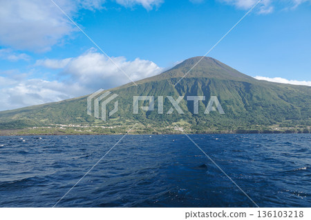 Mount Pico rises above the Atlantic Ocean with green volcanic slopes, textured ridges, and deep blue water under clear daylight 136103218