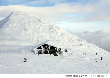 a snow-buried alpine mountain hut on an open ski slope, with snowboarders and skiers passing by 136103803