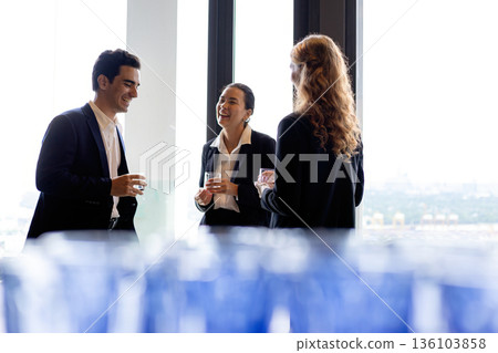 Three business professionals laughing and talking near large office window holding drinks showing friendly networking teamwork confidence and modern corporate culture Three business professionals laughing and talking near large office window holding drinks showing friendly networking teamwork confidence and modern corporate culture 136103858